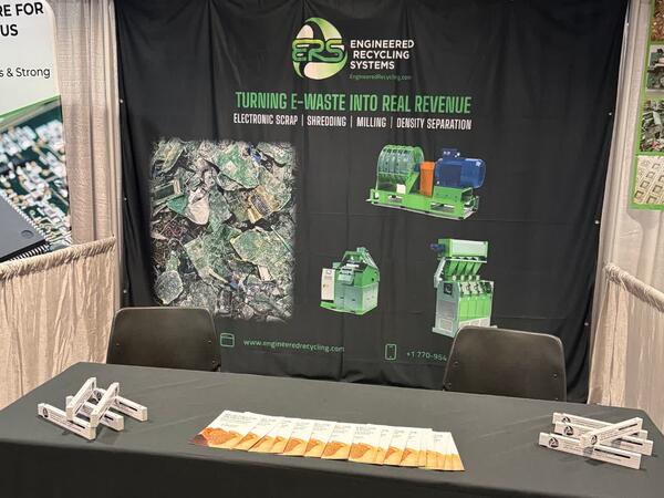 The ERS booth at the E-Scrap Conference in New Orleans, with the black ERS banner reading "Turning e-waste into real revenue — electronic scrap, shredding, milling, density separation" above a table in an ERS cloth, laid out with literature and giveaways.
