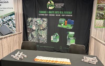 The ERS booth at the E-Scrap Conference in New Orleans, with the black ERS banner reading "Turning e-waste into real revenue — electronic scrap, shredding, milling, density separation" above a table in an ERS cloth, laid out with literature and giveaways.