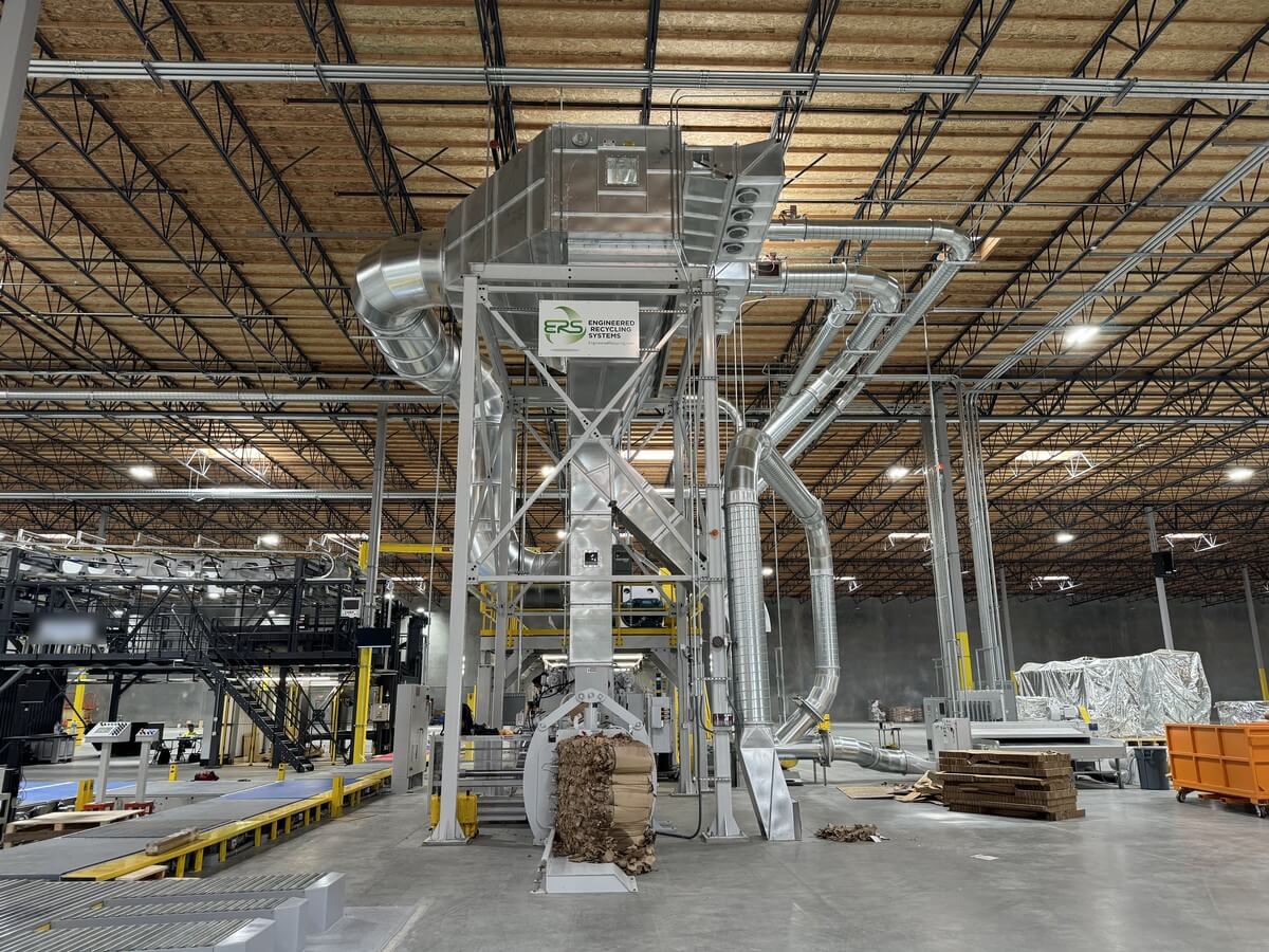 A low profile separator on its tower in a corrugated converting plant, with ERS signage, sweeping duct runs across the hall, collected scrap at the base and the converting line behind